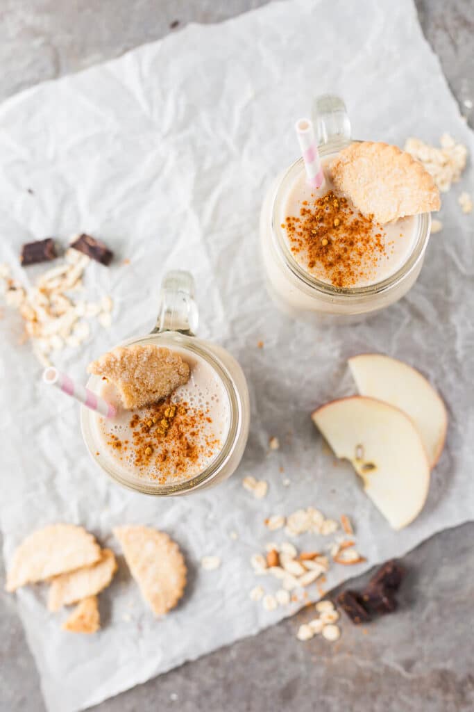A top down picture of two jars full of apple banana smoothie. Cookies, chocolate crumbles and apple slices are spread around the jars.