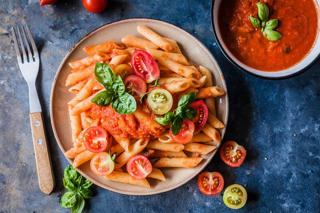 A top down picture of pasta with tomato sauce on a plate. It is garnished with fresh tomatoes cut in half and some basil leaves.