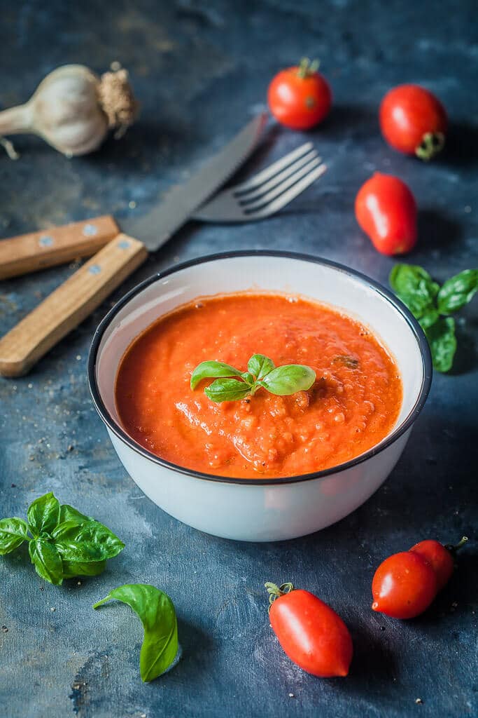 Tomato sauce in a small cup, topped with basil leaves. Tomatoes and basil leaves are spread around the cup.