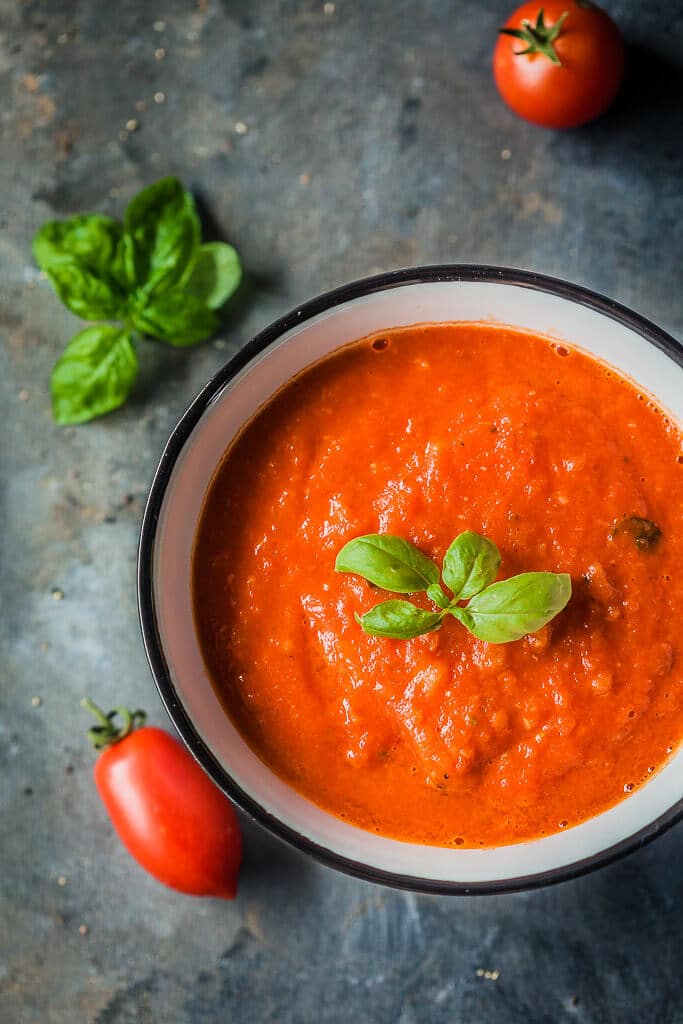 Tomato sauce in a small cup, pictured top down. There are small, fresh tomatoes and basil leaves spread around the cup.