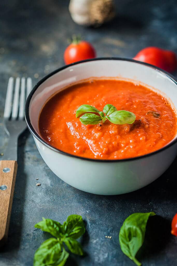 Tomato sauce in a small cup, topped with basil leaves. Tomatoes, garlic, and basil leaves are spread around the cup.