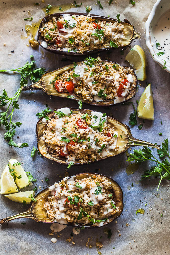 Eggplants stuffed with couscous and topped by lemon tahini sauce. Fresh parsley and lemon slices are spread around them.