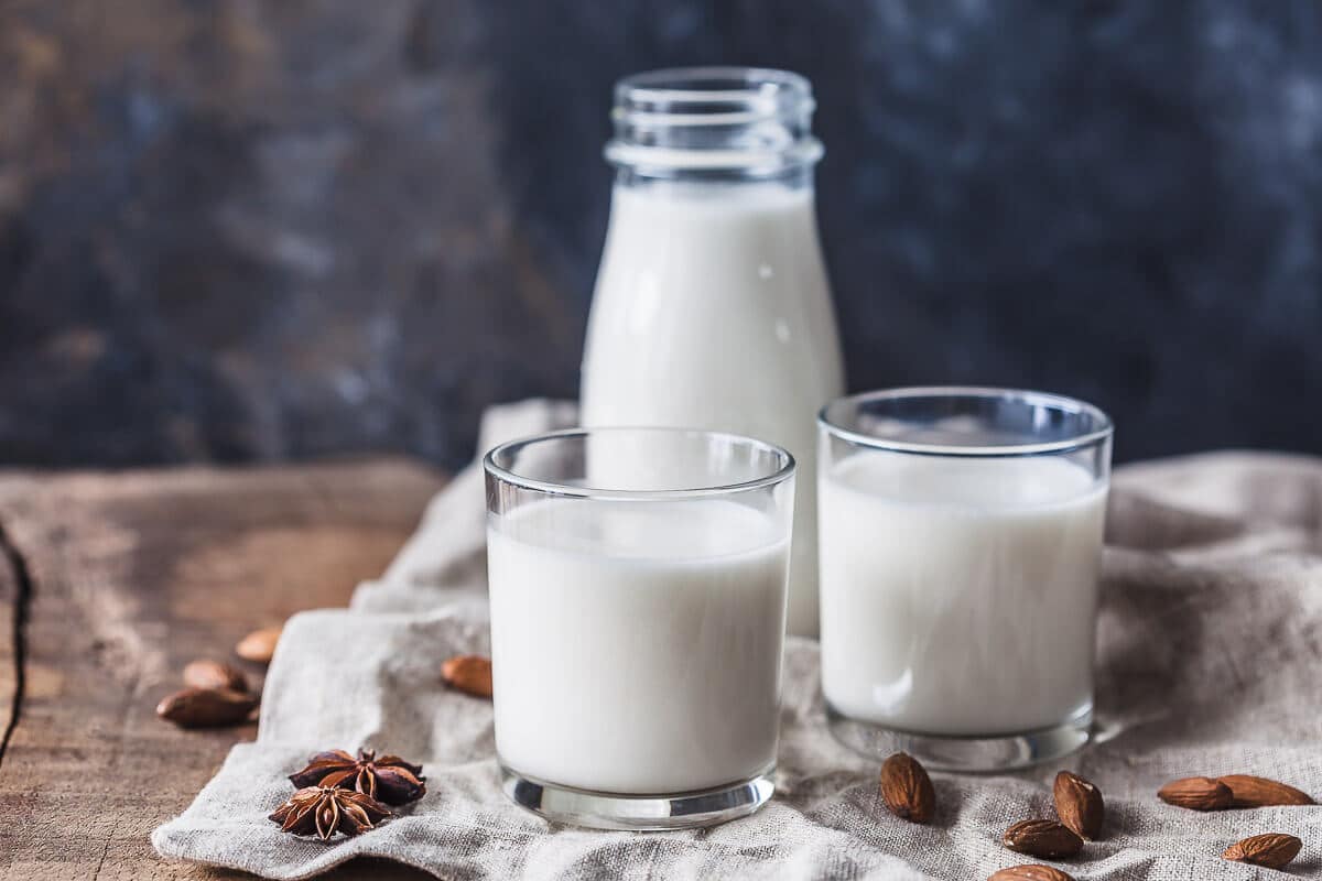 A bottle of almond milk and two glasses of almond milk pictured from the side. There are some almond spread around the glasses.