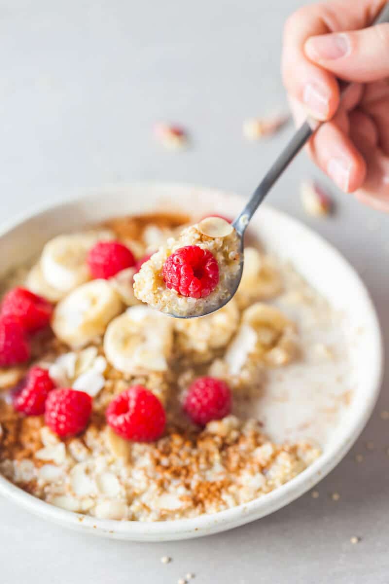 Hand is holding a spoon full of quinoa with a raspberry on top. A bowl of quinoa is visible in the background.